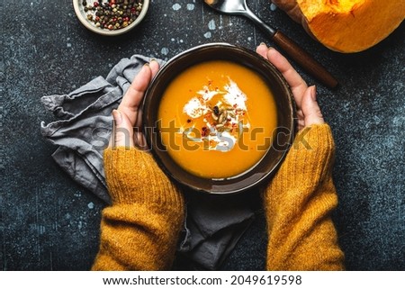 Female hands in yellow knitted sweater holding a bowl with pumpkin cream soup on dark stone background with spoon decorated with cut fresh pumpkin, top view. Autumn cozy dinner concept 
