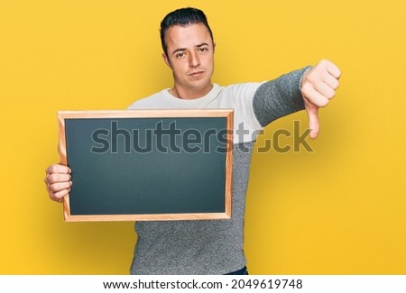 Handsome young man holding blackboard with angry face, negative sign showing dislike with thumbs down, rejection concept 