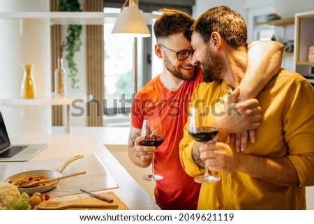 Happy hipster gay couple in love hugging while standing in the kitchen. They are cooking dinner together at home and drinking wine Royalty-Free Stock Photo #2049621119
