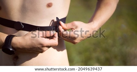 A young male athlete puts on a heart rate monitor and smart watch before jogging in nature. A man monitors his pulse and is engaged in a healthy running at sunset.