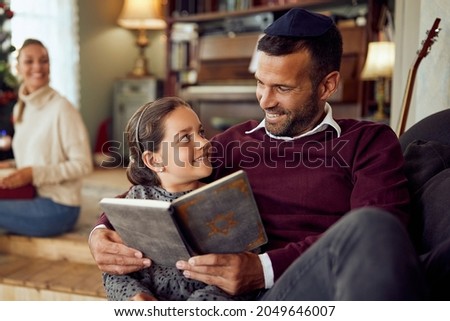 Happy Jewish father and daughter talking while reading Hebrew bible during Hanukkah at home. Royalty-Free Stock Photo #2049646007