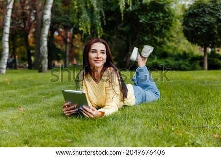 The girl sits in the park and is engaged with the tablet. young brunette sits on the grass and looks into the tablet. girl in the autumn park.