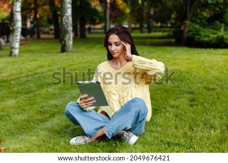 The girl sits in the park and is engaged with the tablet. young brunette sits on the grass and looks into the tablet. girl in the autumn park.