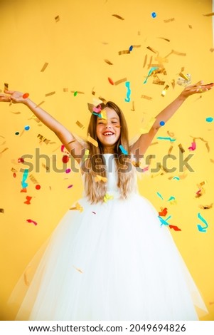 happy little girl with blond hair and in a white dress catches confetti on a yellow background, holiday concept