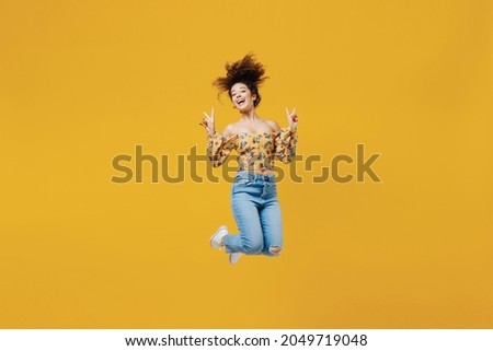 Full length young happy smiling fun woman 20s with culry hair in casual clothes jumping high show victory sign gesture isolated on plain yellow background studio portrait. People lifestyle concept