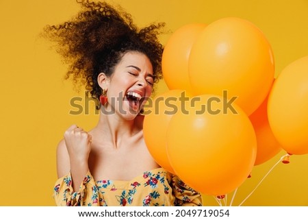 Young happy woman with culry hair in casual clothes celebrating birthday holiday party hold bunch of colorful air inflated helium balloons do winner gesture isolated on plain yellow background studio