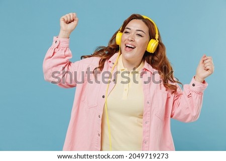 Young fun smiling caucasian redhead chubby overweight woman 30s wear pink shirt casual clothes wireless yellow headphones listen to music dancing isolated on pastel blue background studio portrait.