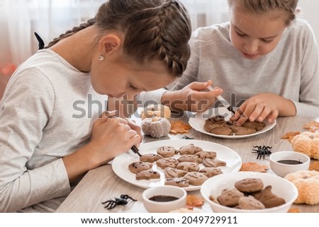 Two girls decorate halloween gingerbread cookies on plates with chocolate icing. Cooking treats for halloween celebration. Lifestyle Royalty-Free Stock Photo #2049729911