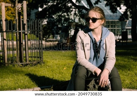 Girl in sunglasses and light coat is sitting in the park at sunny autumn day.