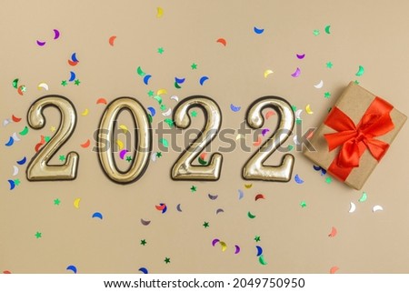 Top view on beige background of gold digits 2022 with red bow gift box decorated with multicolored sequins. Concept, layout of a New Year's card, packaging.
