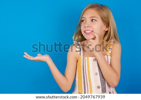 Positive Caucasian blonde kid girl wearing dress against blue wall advert promo touch finger teeth