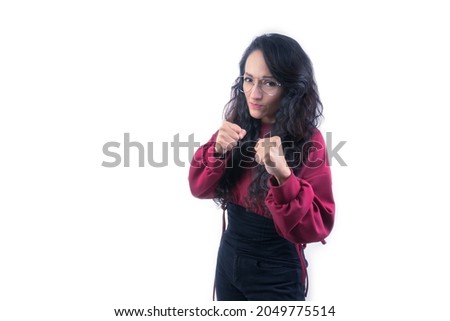 Pretty latin hispanic woman with long thick hair wearing glasses on white background indoors doing expressions and gestures, showing fists, in fighting pose, excited, upset, furious