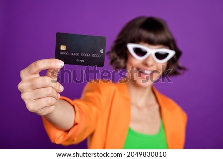 Photo of pretty excited young woman dressed orange blazer dark eyewear showing debit card smiling isolated purple color background