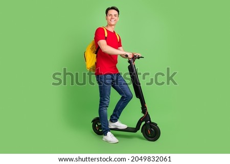 Photo of pretty charming young guy dressed red t-shirt driving kick scooter smiling isolated green color background