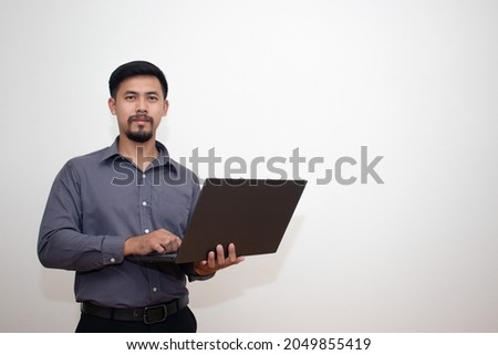 Portrait of a young modern businessman standing holding a laptop and looking at the camera with a happy smile at his work. isolated on a gray background