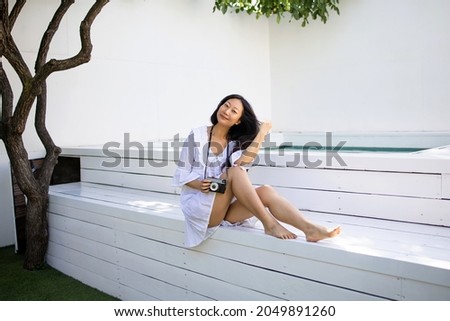 Positive asian woman in trendy white dress relaxing near swimming pool of modern hotel and taking pictures with retro camera
