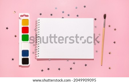 Empty notebook with paints, brush and confetti stars pours on a pastel pink background, top view. Flat lay. Holiday, greeting or good news concept. Mockup template. top view. Blank notepad