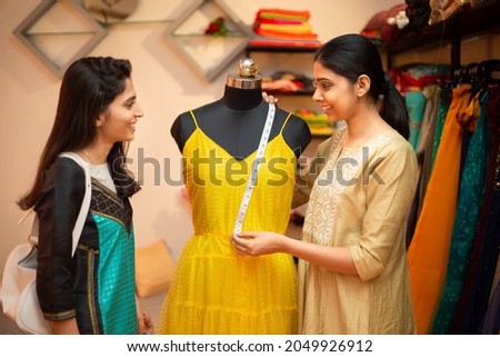 Young happy indian woman,tailor seamstress,smiling fashion designer showing customer or client dress measurements with tape,measuring on mannequin,standing in garment workshop,atelier or studio. Royalty-Free Stock Photo #2049926912