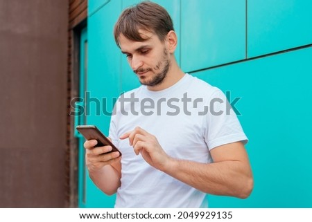 A young man in a white blank T-shirt stands against the background of a blue wall and calls on the phone. The guy is holding a smartphone. Mock-up.