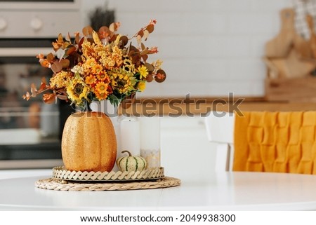 Still-life. Yellow, orange flowers in a vase, pumpkins and candles on a golden tray on a white table in a home kitchen interior. A cozy autumn concept. Royalty-Free Stock Photo #2049938300