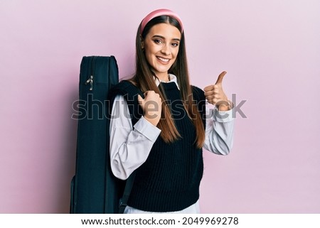 Beautiful brunette young musician woman wearing guitar case smiling happy and positive, thumb up doing excellent and approval sign 