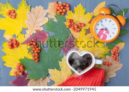 A white mug in the shape of a heart with steaming coffee or tea, an orange alarm clock and a red scarf on leaves.