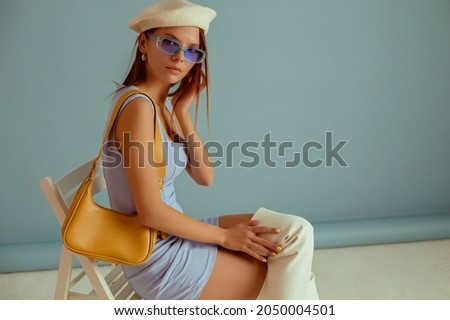 Fashionable woman wearing classic white beret, color sunglasses, elegant mini dress, over knee boots, with yellow leather shoulder bag, posing on blue background. Copy, empty space for text Royalty-Free Stock Photo #2050004501