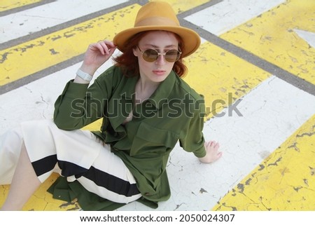 Sporty ginger girl wearing fashion sport chic clothes sitting on street zebra crossing road, outdoor shoot, urban style. Female model in swag clothes posing outside over pedestrian crosswalk.
