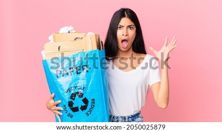 young pretty hispanic woman screaming with hands up in the air and holding a paper bag to recycle