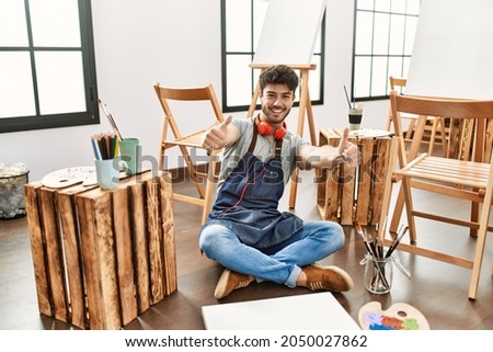 Young hispanic man sitting at art studio approving doing positive gesture with hand, thumbs up smiling and happy for success. winner gesture. 