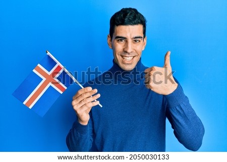 Handsome hispanic man holding island flag smiling happy and positive, thumb up doing excellent and approval sign 
