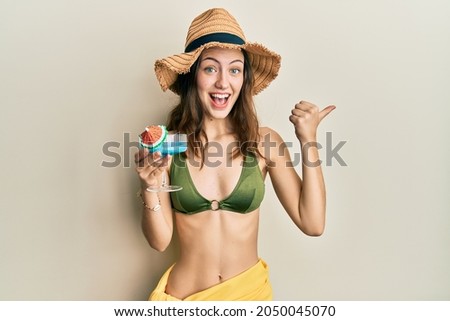 Young brunette woman wearing bikini drinking cocktail pointing thumb up to the side smiling happy with open mouth 