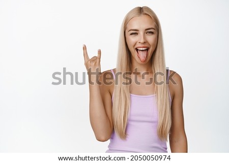 Sassy blond girl showing rock-n-roll, heavy metal horns and tongue, enjoying event, having fun, standing over white background