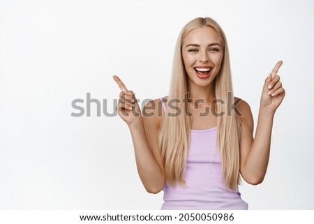 Portrait of beautiful blond woman smiling, pointing fingers sideways, showing left and right variants, giving you options, standing over white background