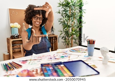 Beautiful african american woman with afro hair painting at art studio smiling making frame with hands and fingers with happy face. creativity and photography concept. 