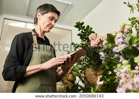 Low angle portrait of adult female florist doing stock inventory while working in flower shop, small business concept