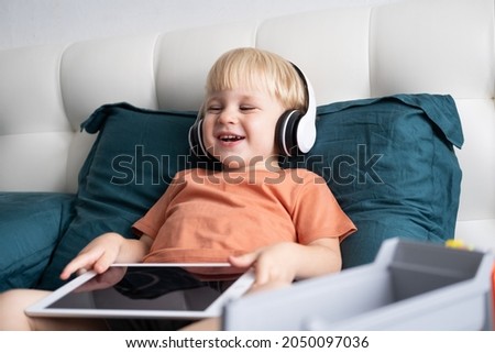 Smiling happy little kid boy using tablet in wireless headphones in bed at home.