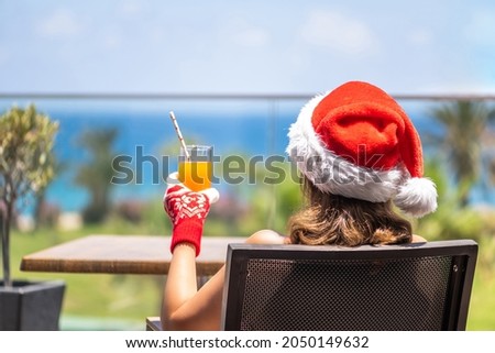 Woman in christmas santa claus hat relaxing and drinking cocktail on balсony opposite palm trees beach and sea. New year