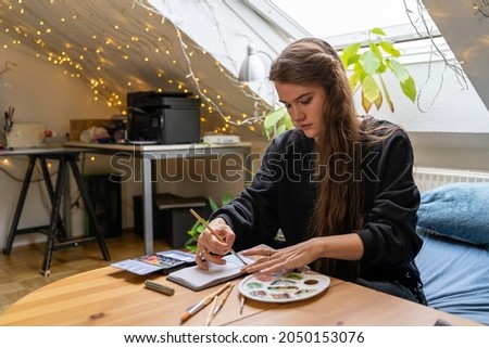 Young brunette woman student drawing nature landscape in paper notebook with colorful watercolor palettes and brushes in dormitory bedroom, sitting on bed near table and pumpkin on autumn weekend day