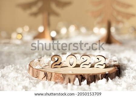 New year 2022. Numbers 2022 on wooden stand on beige pastel blurred background with decorative fir trees, snow and lights. Christmas greeting card.