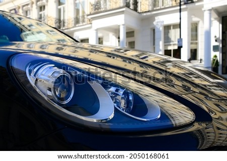 White facade of a typical old London town building reflected on the bonnet or engine hood of dark blue sports car parking in front of the renovated house. Distorted warped mirror image on varnish