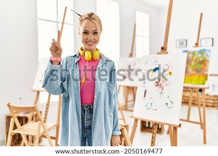 Young caucasian girl at art studio showing and pointing up with finger number one while smiling confident and happy. 