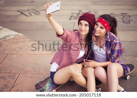 Two Brunette teenage girls friends in hipster outfit (jeans shorts, gumshoes, plaid shirt, hat) with a skateboard at the park outdoors make selfie on a phone. Copy space