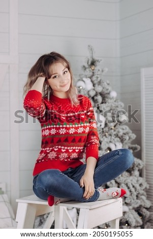 A young caucasian charming smiling woman in a red Christmas sweater with deer and snowflakes pattern siitting on the background of a white fir tree. New Year's interior