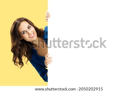 Happy smiling brunette young woman in blue clothing, standing behind, peeping from blank banner billboard or mock up signboard with copy space for some ad text, isolated over yellow background. Female