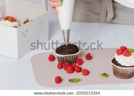 A woman prepares a cake with raspberries and white cream. Women's hands spread cream on a cupcake and decorate it with berries. Homemade dessert.