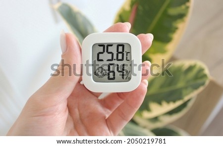 Thermometer hygrometer in hand against the background of a plant for which good humidity and temperature are important.  A hand holds a thermometer a hygrometer that shows the humidity and temperature Royalty-Free Stock Photo #2050219781