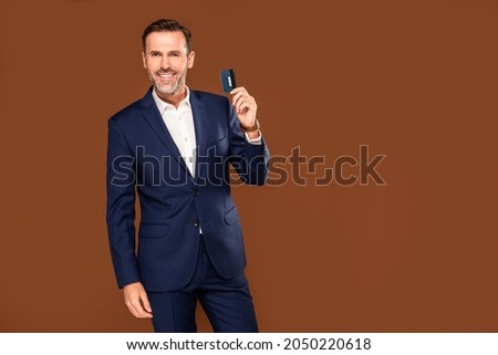 Photo of handsome happy businessman holding credit card , posing over brown studio background, looking at the camera. 
