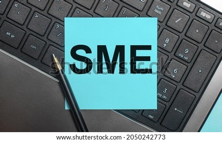 The computer keyboard has a pen and a blue sticker with the text SME SUBJECT MATTER EXPERT. Flat lay.