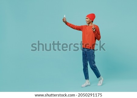 Full body young happy african american man 20s in orange shirt hat doing selfie shot on mobile cell phone post photo on social network isolated on plain pastel light blue background studio portrait.
